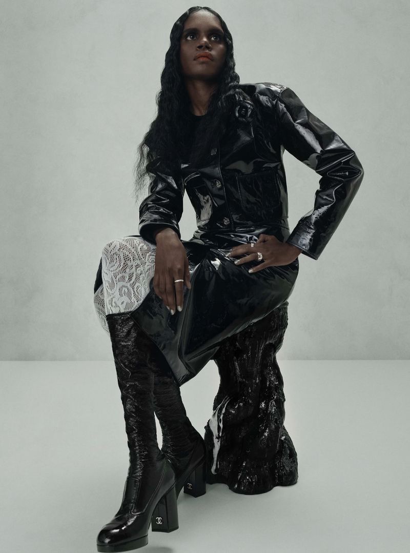 A woman in a black shiny coat and boots poses while sitting on a black sculptural object against a neutral background in a studio shot.