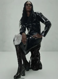 Model wearing a shiny black coat and heeled boots seated on a dark textured stool against a neutral backdrop