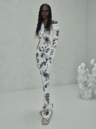 Model wearing a white floral long-sleeve dress and matching gloves standing in a minimalist studio setting.