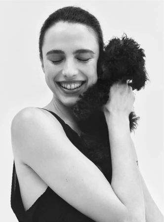 Margaret Qualley smiling with eyes closed while holding a small black puppy against her chest on a white background