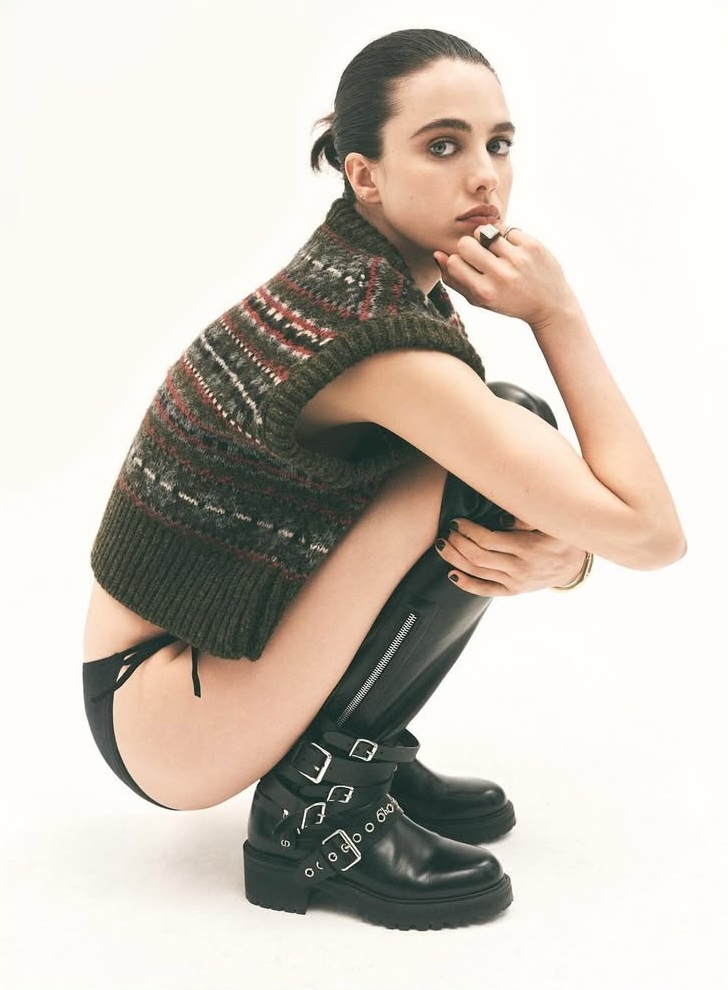 A young woman with dark hair squats in a studio, wearing a patterned vest, dark shorts, and black platform boots with buckle details.