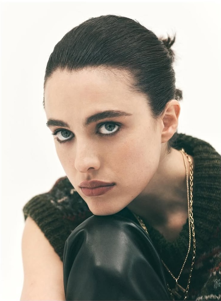 Close-up of a young adult woman with dark hair and light eyes, wearing a dark green patterned sweater vest and gold necklaces in a studio.