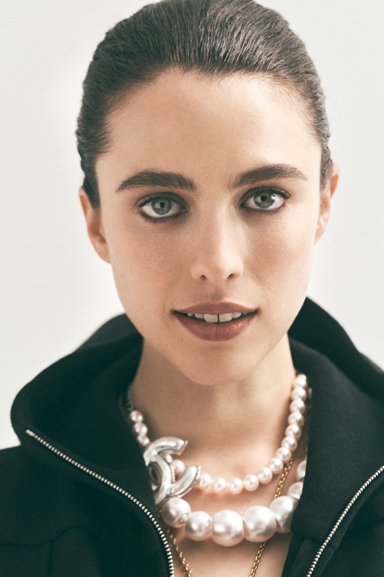 Close-up portrait of a woman with slicked-back hair wearing a pearl necklace with a Chanel logo against a clean, bright background.