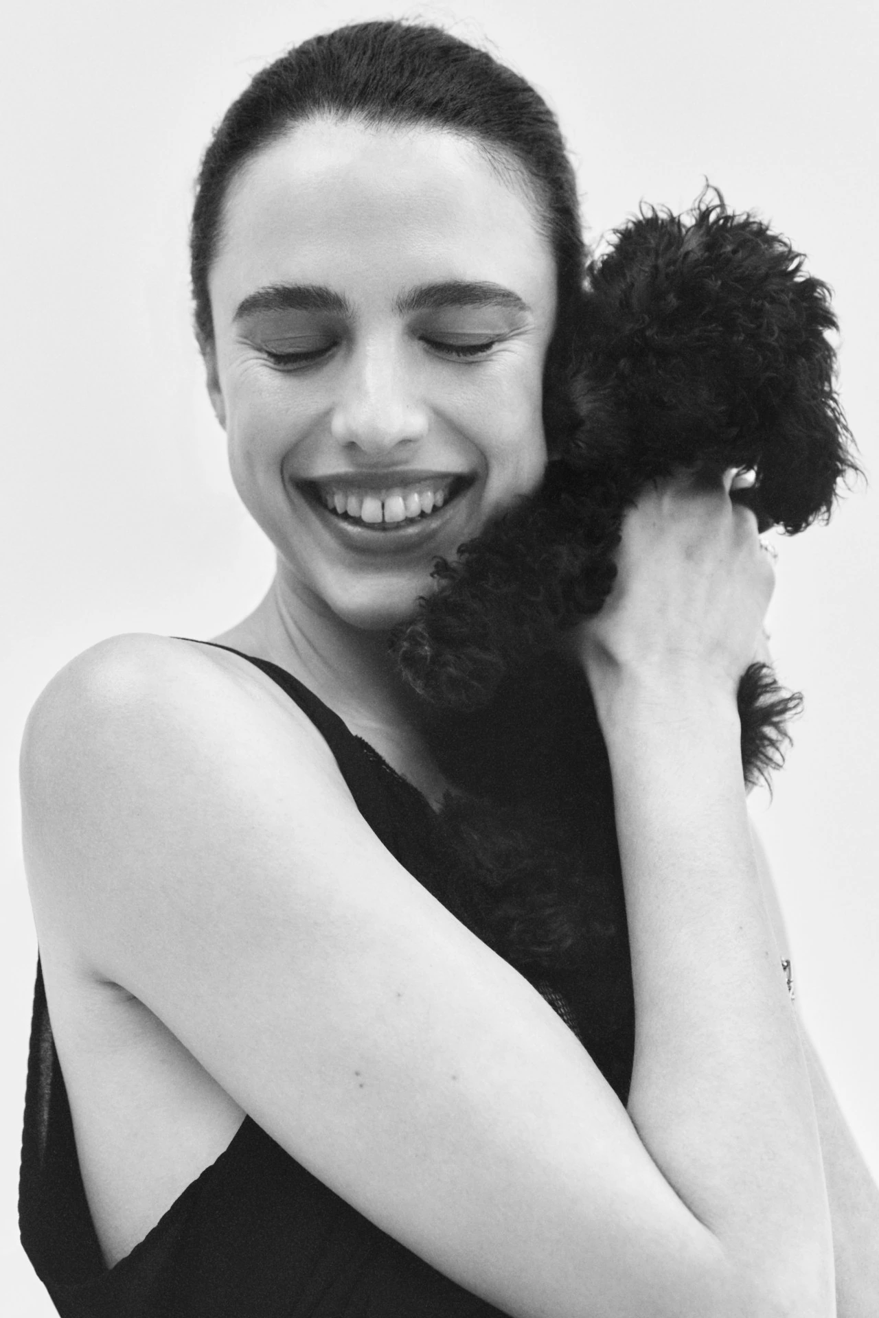 Portrait of a smiling woman in a black top hugging a black miniature poodle against a bright white background, creating a soft, intimate feel.