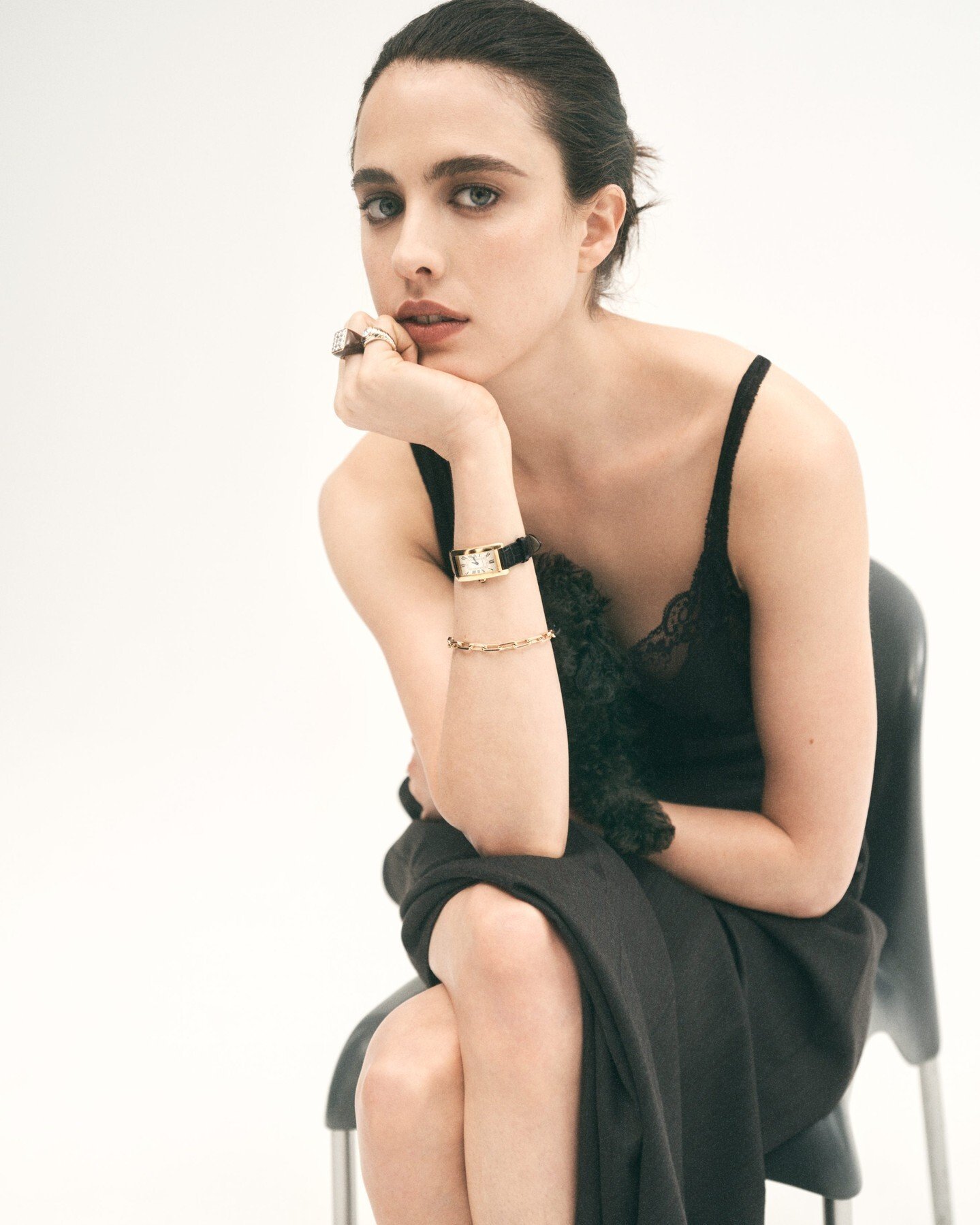 Elegant woman with black hair and dress sits indoors; she has her hand to her face with a ring on and is wearing a watch.