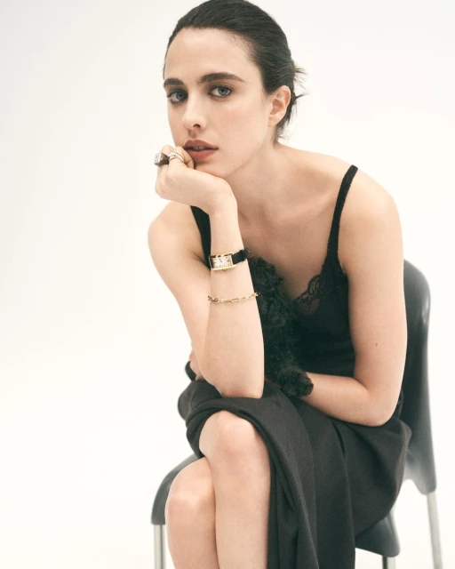 Elegant woman with black hair and dress sits indoors; she has her hand to her face with a ring on and is wearing a watch.