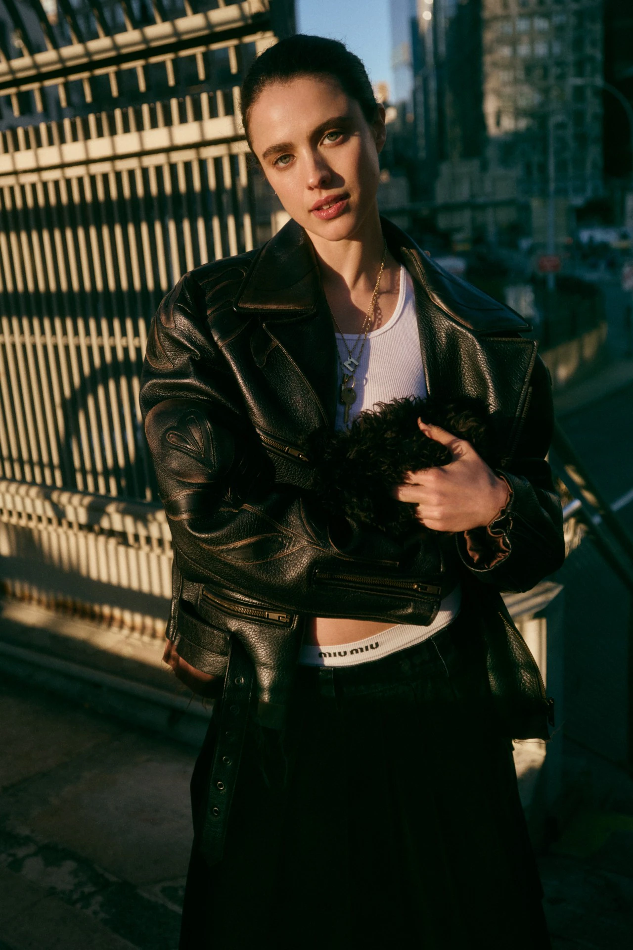 Margaret Qualley wearing a black leather jacket and white tank top outdoors against an urban background with warm sunlight.