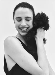 Margaret Qualley smiling with eyes closed while holding a small black puppy against her chest on a white background