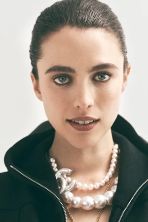 Close-up portrait of Margaret Qualley wearing a dark zipped jacket and layered pearl necklaces against a white studio background.