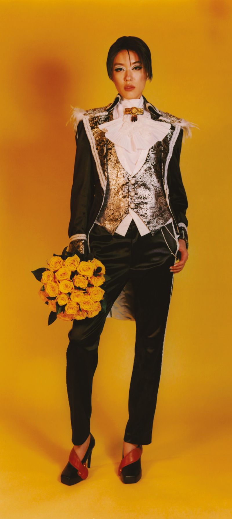 Full-length portrait of a woman in a decorative tuxedo holding yellow roses, set against a bright yellow backdrop.