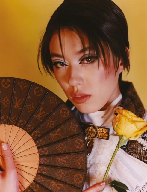 Close-up of a woman holding a Louis Vuitton fan and yellow rose. She is wearing a white top, brown jacket and has heavy white eye shadow.