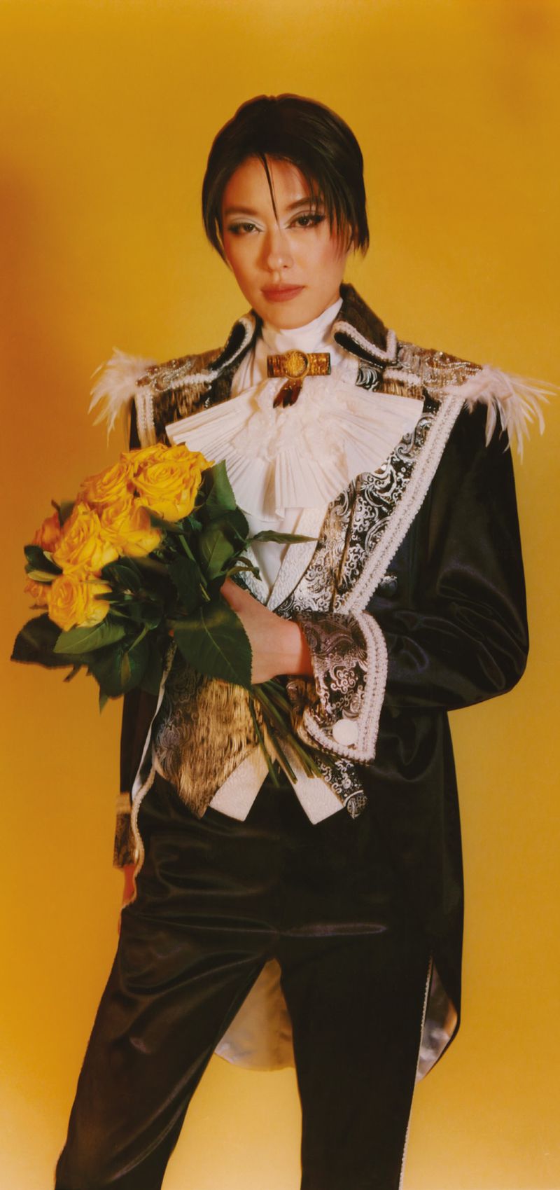 Portrait of an androgynous person holding a bouquet of yellow roses, wearing a black and gold decorated jacket against a yellow backdrop.