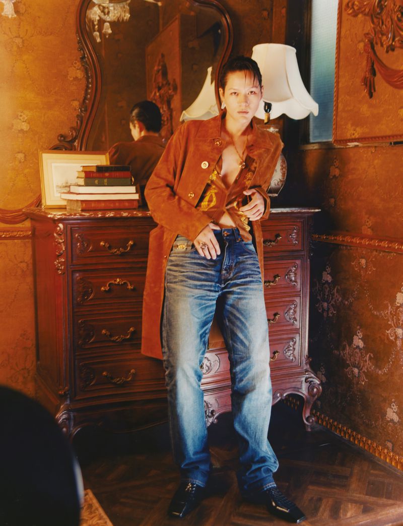 A woman poses in an orange coat and jeans, leaning against a vintage dresser in a room with patterned wallpaper and a large mirror.