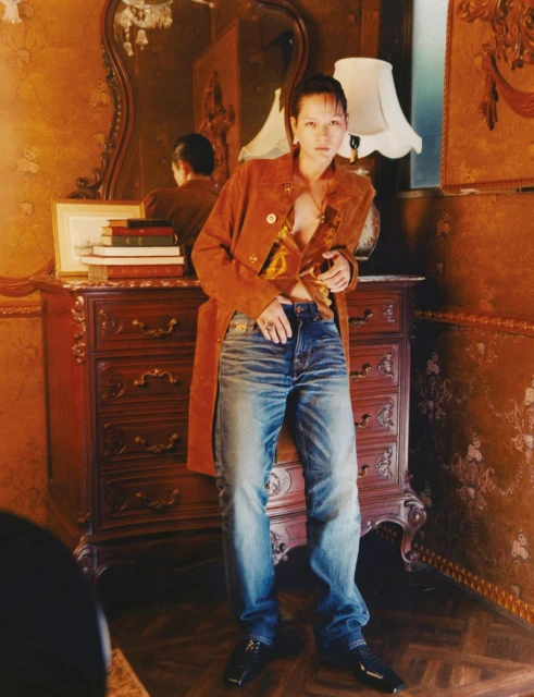 A woman poses in an orange coat and jeans, leaning against a vintage dresser in a room with patterned wallpaper and a large mirror.