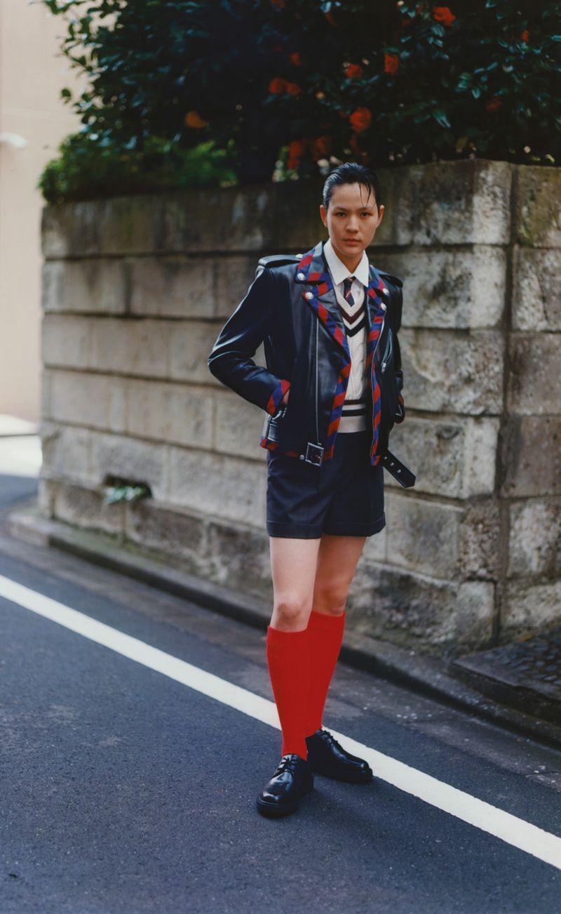 Fashionable woman in a moto jacket and skirt standing on a street, posing confidently with red socks and shiny black shoes in urban setting.