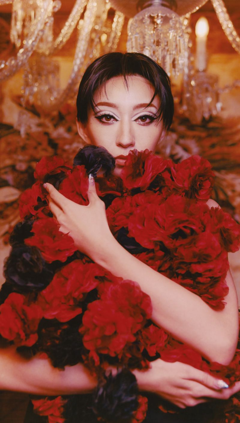 Adult woman with dark hair, wearing a red and black floral garment in an indoor environment with a chandelier background.