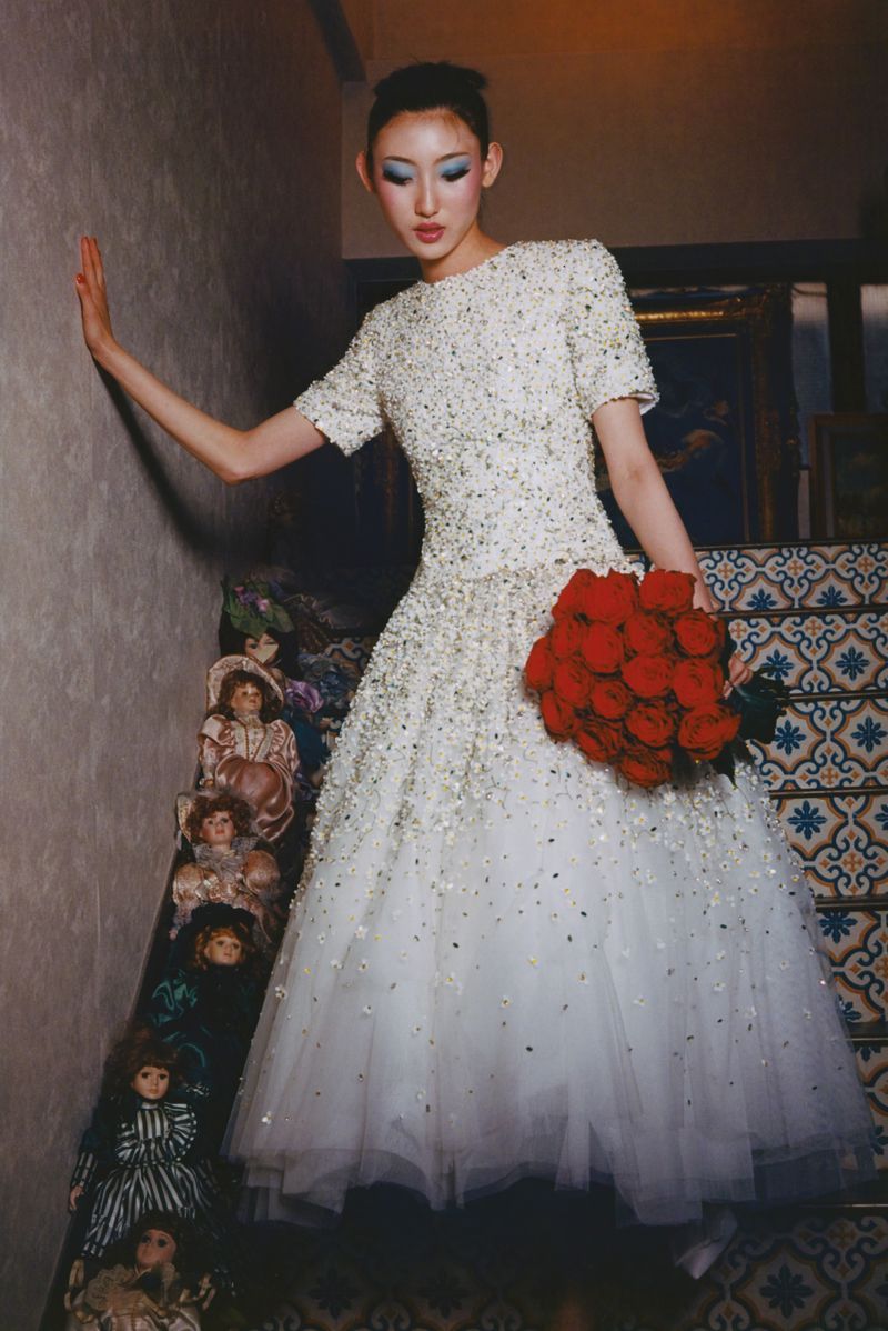 An Asian woman poses in a white dress with a red rose bouquet on stairs lined with antique dolls, creating a surreal fashion portrait.