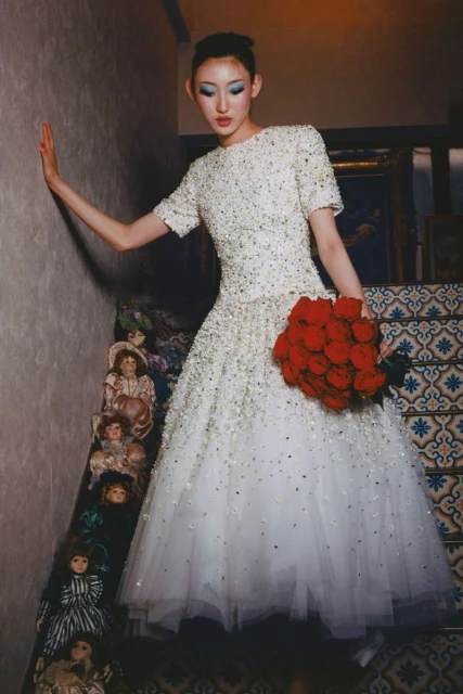 An Asian woman poses in a white dress with a red rose bouquet on stairs lined with antique dolls, creating a surreal fashion portrait.