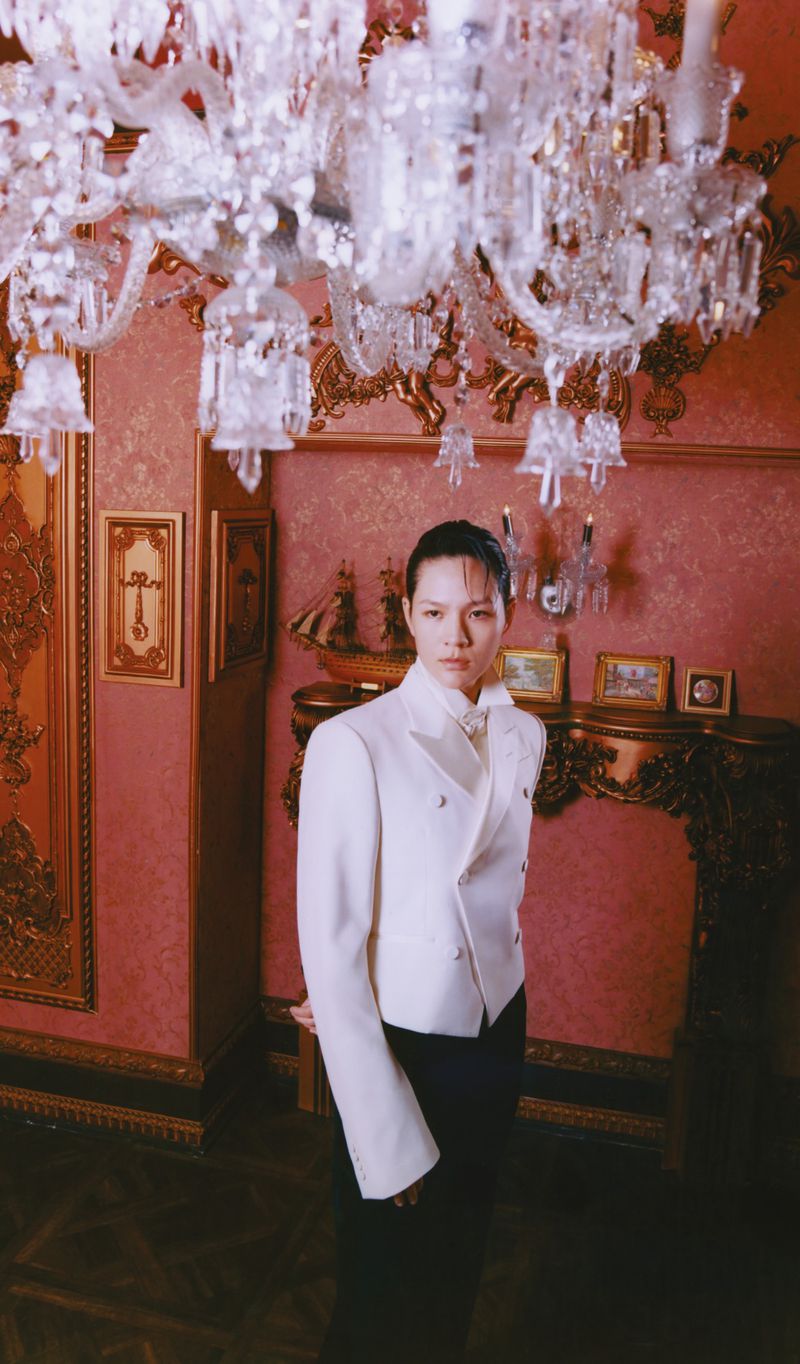 Elegant woman in a white suit poses indoors beneath a crystal chandelier, with an ornate room featuring gold trim and framed pictures.