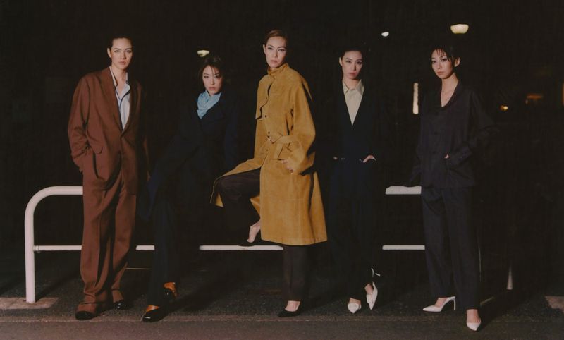 Five women in dark suits and coats pose against a dark background, illuminated by soft light, showcasing fashion and style.