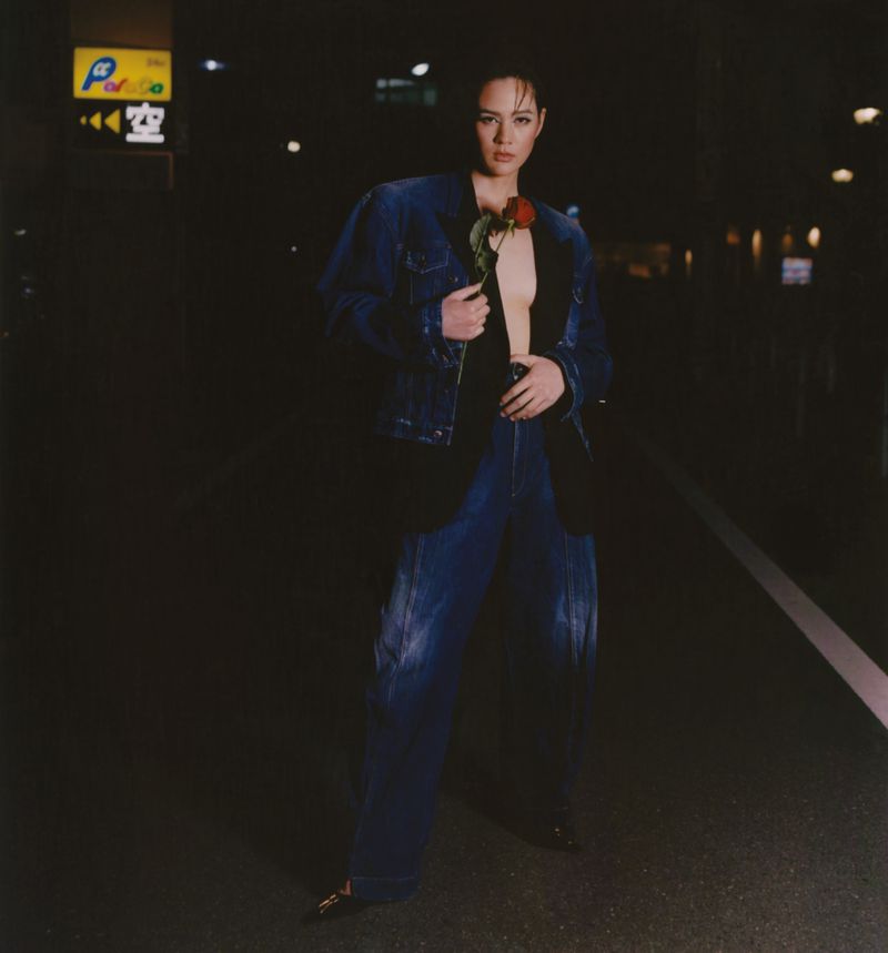 Woman in denim jacket and pants poses on a city street at night, holding a red rose with a neon sign in the background.