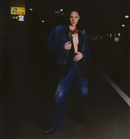 Woman in denim jacket and pants poses on a city street at night, holding a red rose with a neon sign in the background.