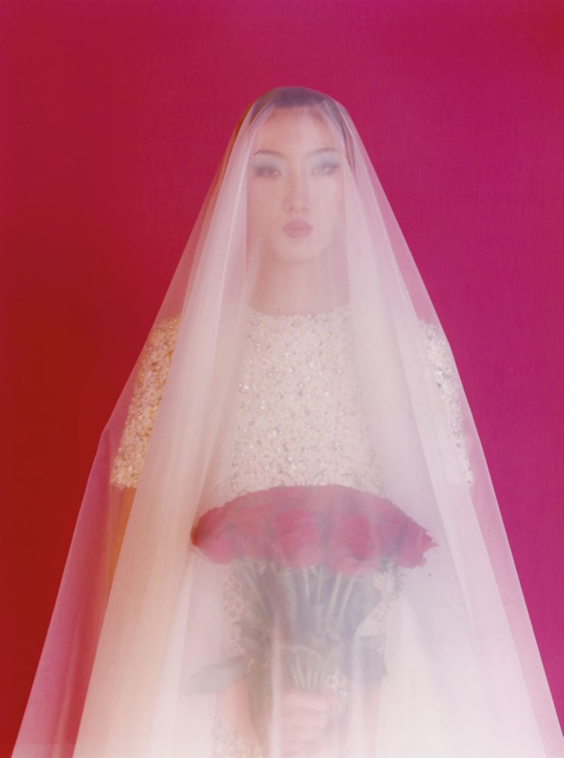 Elegant Asian woman in an embellished top and veil holds a bouquet of roses against a magenta backdrop in a soft studio portrait.