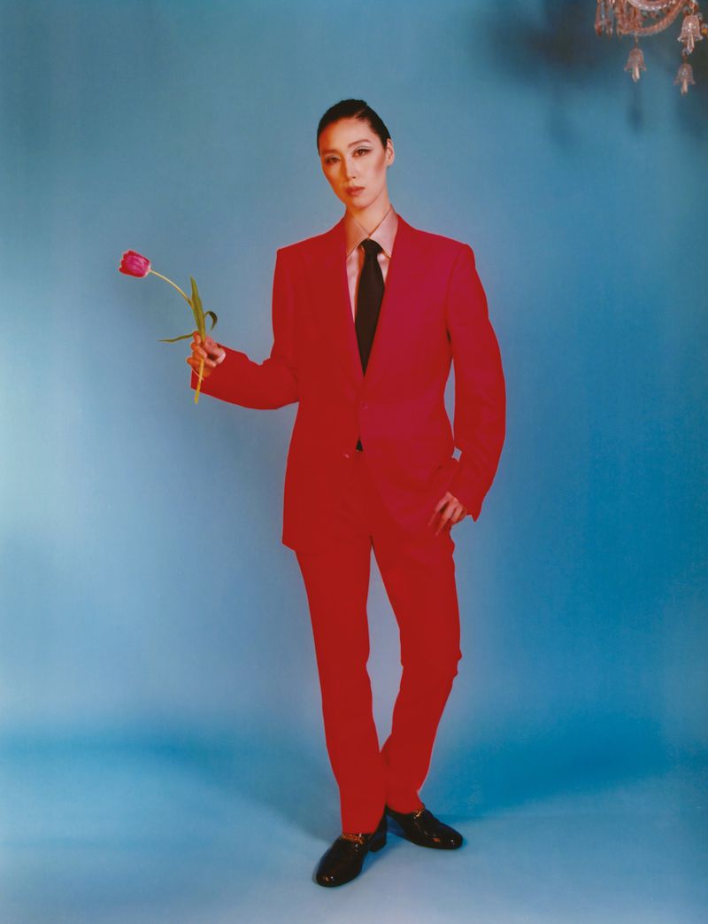 Portrait of an Asian woman in a vibrant red suit, holding a tulip against a light blue backdrop with a vintage chandelier.