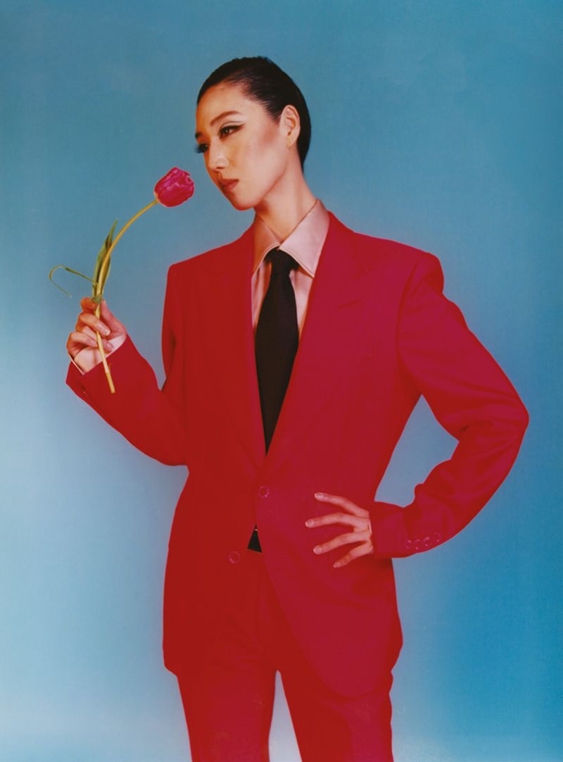 A woman in a red suit poses with a red tulip against a blue background, creating a striking fashion or beauty portrait.