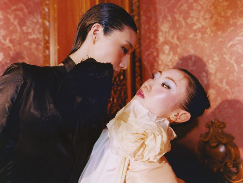 Two Asian women pose in a retro-style studio photograph. One is in black and the other in white, with bold makeup.