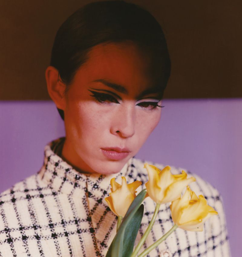 Portrait of a woman with dramatic eyeliner holding yellow tulips in front of a purple and brown backdrop, captured in a close-up shot.