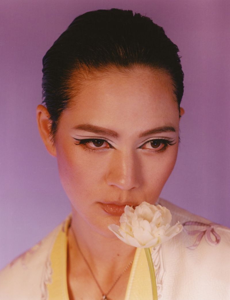 Portrait of an adult female with dramatic eye makeup holding a white tulip against a soft lavender background in a close-up shot.