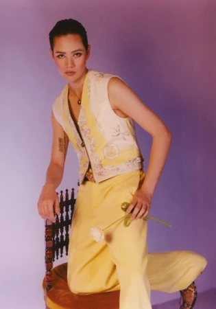 Adult female in a yellow pantsuit sitting on a velvet chair, holding a white rose in a studio setting with a purple background.