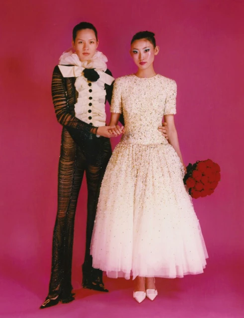 Two people, one in a white dress with red flowers, and one in a black and white suit, stand in front of a pink backdrop.