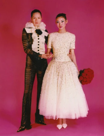 Two people, one in a white dress with red flowers, and one in a black and white suit, stand in front of a pink backdrop.