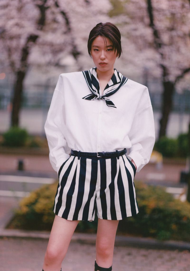 Fashion shot of an Asian woman in a sailor-style white blouse and black-and-white striped shorts, posing in an urban park with flowering trees.