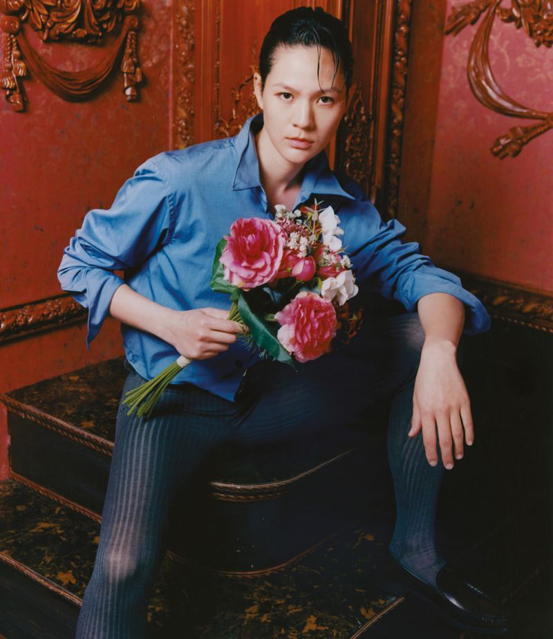 Elegant woman with slicked back hair poses on steps, holding bouquet. She wears a blue shirt and dark striped leggings in an ornate indoor setting.