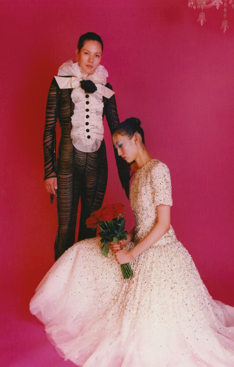 Two women in elaborate couture poses against a pink backdrop, one seated in a gown holding roses, the other standing in a ruffled collar.