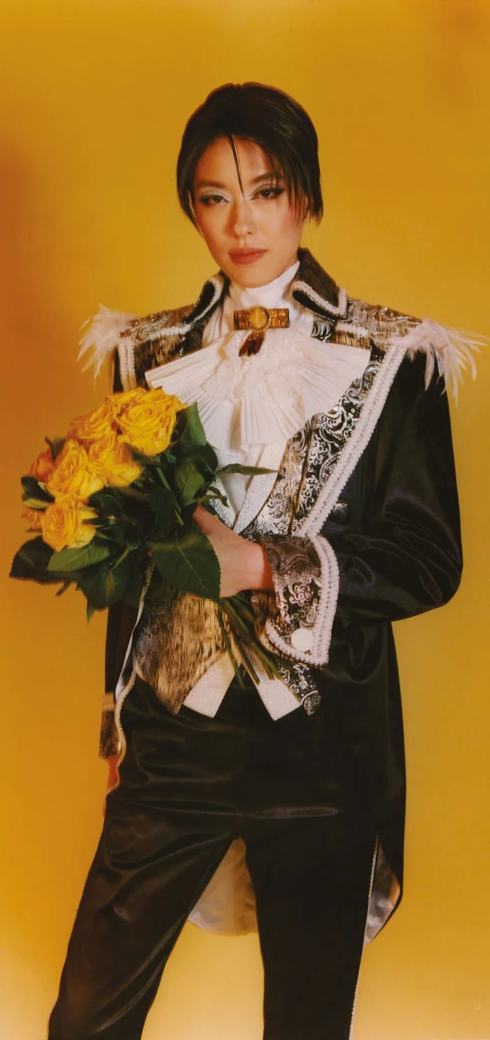 Model in ornate black tailcoat holding yellow roses against a warm yellow background for The Face Magazine.