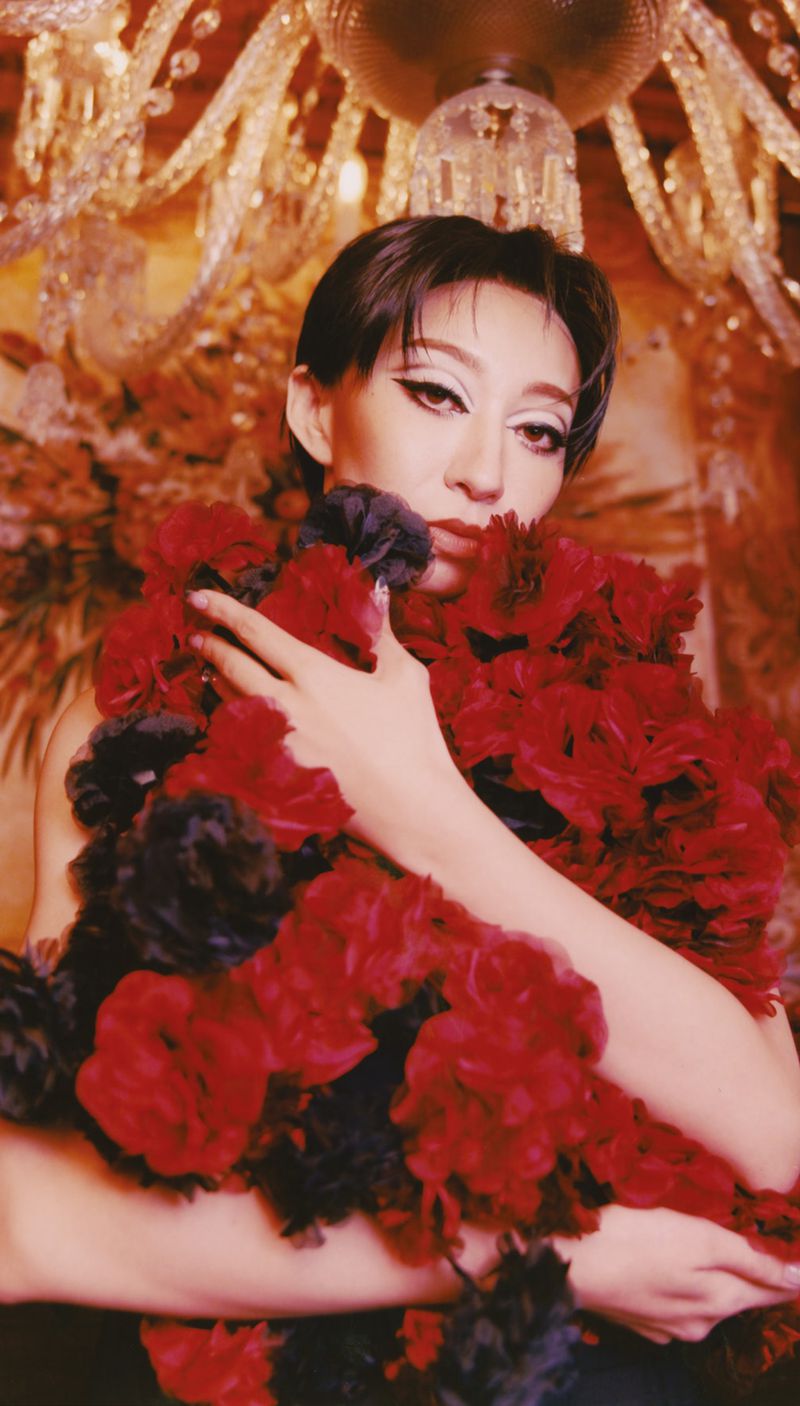 Model holding a large bouquet of deep red flowers beneath an ornate glass chandelier for The Face Magazine.