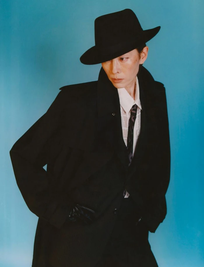 Model in black coat and fedora hat posing against a solid blue background for The Face Magazine editorial