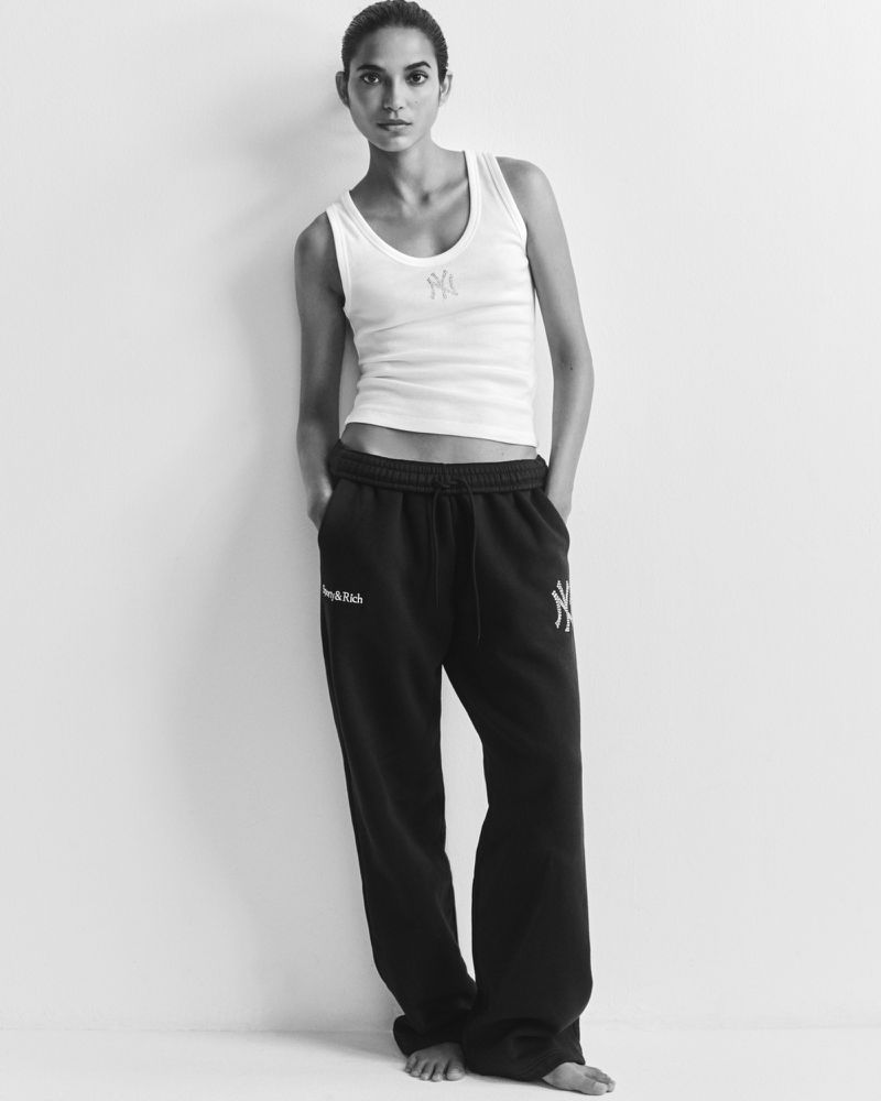 Model Amrit standing barefoot in a white ribbed tank top and black sweatpants against a white studio background.