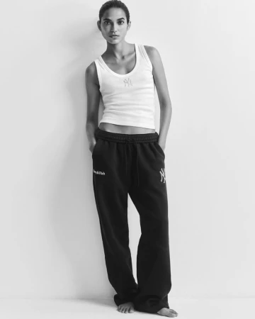 Model Amrit standing barefoot in a white ribbed tank top and black sweatpants against a white studio background.