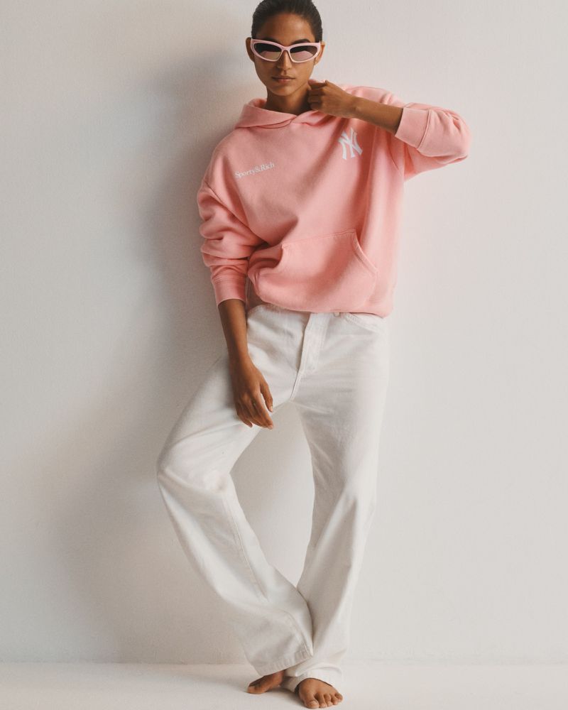 Model wearing a pink hooded sweatshirt, white wide-leg pants, and sunglasses standing against a plain white studio backdrop.