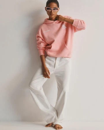 Model wearing a pink hooded sweatshirt, white wide-leg pants, and sunglasses standing against a plain white studio backdrop.
