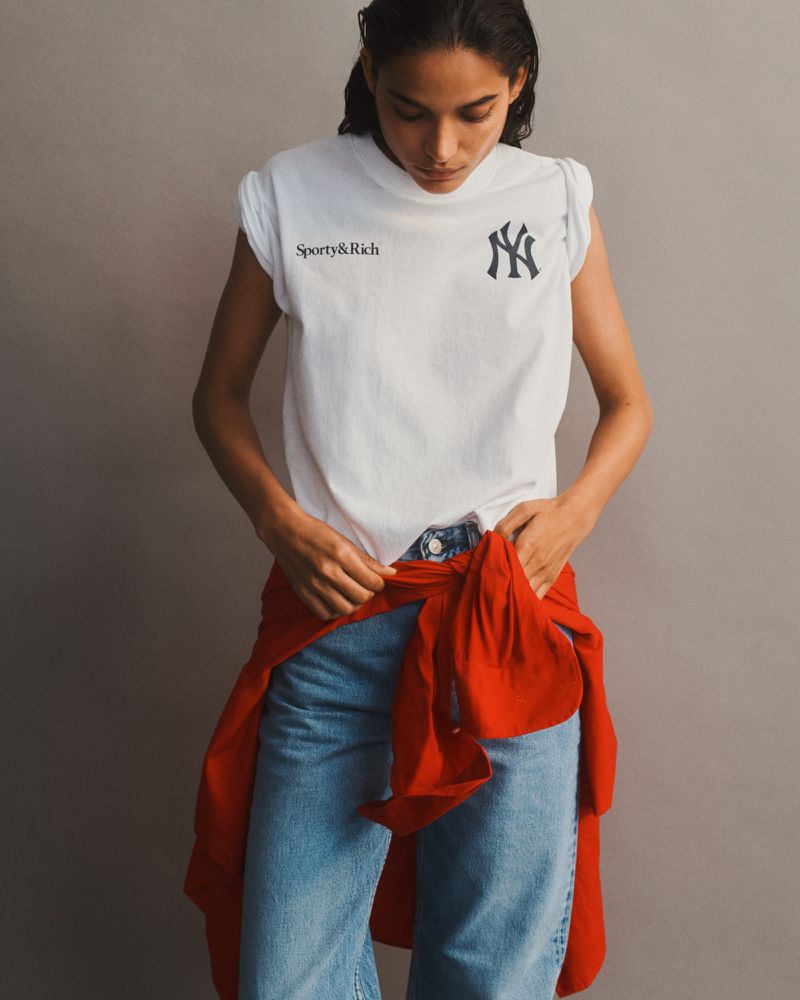 Model Amrit wearing a white Sporty & Rich t-shirt, blue jeans, and a red shirt tied at the waist against a grey studio backdrop.