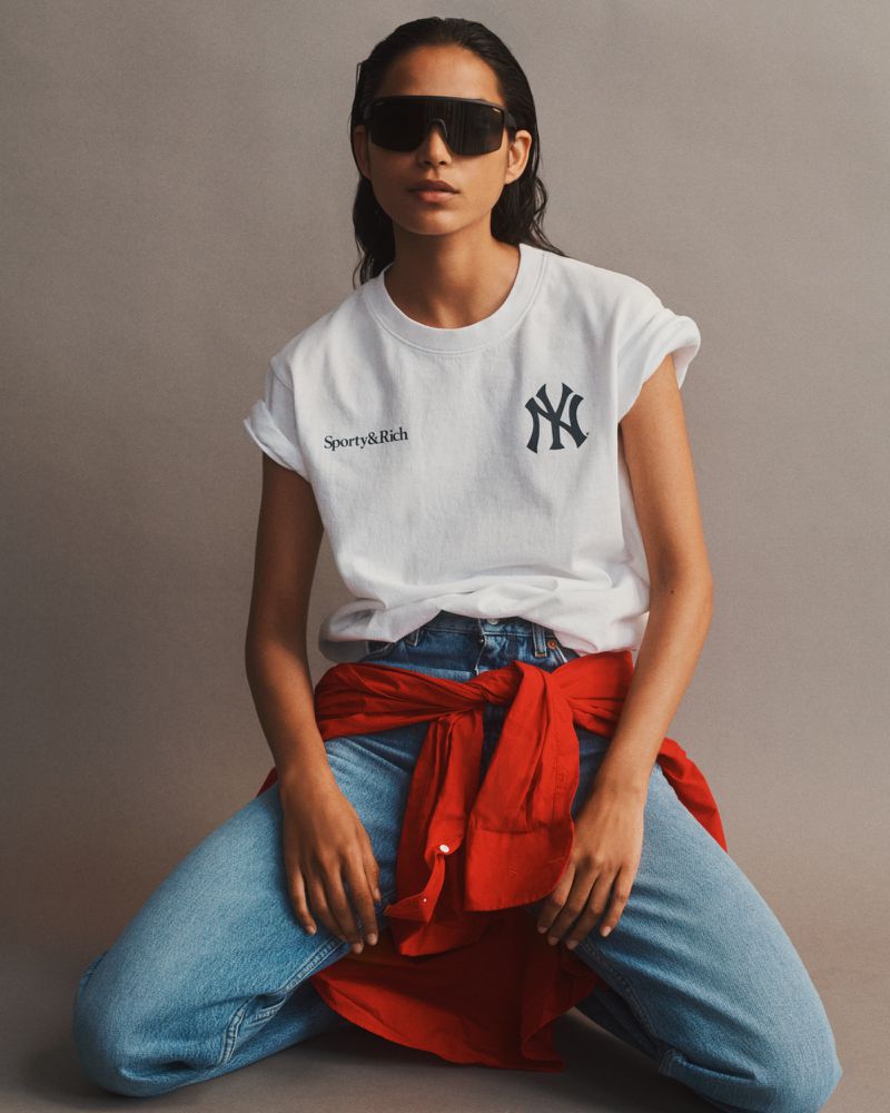 Model Amrit wearing a white Sporty & Rich logo t-shirt, dark shield sunglasses, blue denim jeans, and a red shirt tied around the waist.