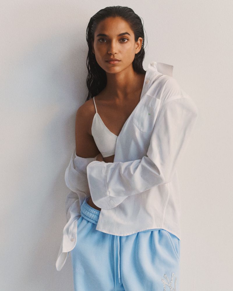 Model Amrit wearing an open white button-up shirt over a white bralette and light blue sweatpants against a white backdrop.