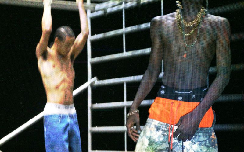 Two shirtless male models posing in streetwear, one wearing layered orange and printed shorts alongside beaded necklaces.