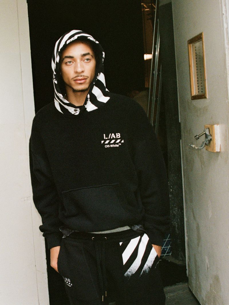 Model Julez Smith wearing a black Off-White hoodie with a striped hood and matching sweatpants standing in an open doorway.
